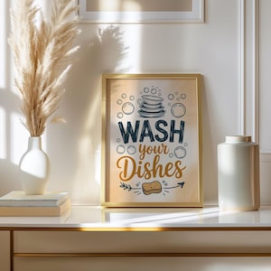 May include: A framed wall art print featuring a stack of plates, bubbles, and a sponge illustration with the text "WASH your Dishes" in dark blue and gold lettering on a light beige background, displayed in a thin gold metal frame.