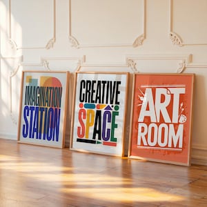 May include: Three framed art prints with the words "IMAGINATION STATION", "CREATIVE SPACE", and "ART ROOM" in bold, colorful fonts. The prints are in wooden frames and lean against a white wall with decorative molding, on a wooden floor.
