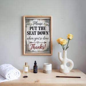 Puede incluir: Un letrero de madera enmarcado con el texto "Please PUT THE SEAT DOWN when you're done Thanks!" se muestra. El letrero está encima de una mesa de madera con una toalla blanca enrollada, un frasco de perfume, una botella oscura, un pequeño recipiente blanco y gris, un jarrón con rosas amarillas y un lápiz de madera.