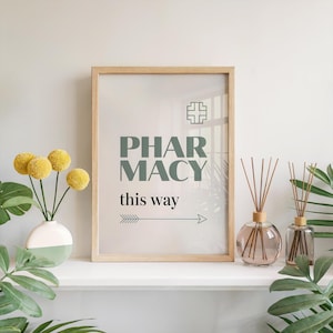 May include: A framed print with the word "PHARMACY" in teal letters, along with the phrase "this way" and an arrow. The frame is light wood. The print is on a white shelf with a vase of yellow flowers and two reed diffusers.