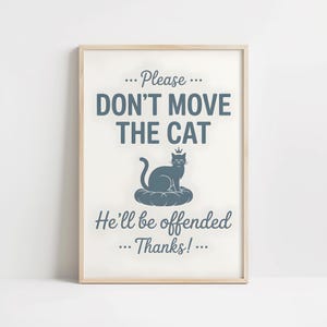 May include: A framed print with a light wood frame. The print features the text "... Please ... DON'T MOVE THE CAT He'll be offended ... Thanks! ..." in a dark teal color. A teal cat wearing a crown is centered on the print.