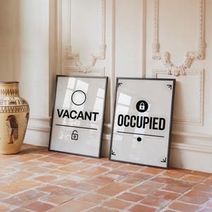 May include: Two framed signs with black text on a white background. One sign reads "VACANT" with a circle and a lock icon. The other reads "OCCUPIED" with a lock icon and a dot. The signs are leaning against a wall.