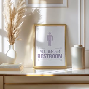 May include: A framed sign with the text "ALL GENDER RESTROOM" in purple, featuring a gender-neutral symbol. The sign is in a gold frame, set on a white surface with a vase of pampas grass and a decorative white vase.
