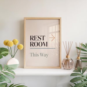 May include: A framed sign with the words "REST ROOM This Way" in a modern font. The sign is in a light wood frame and is accompanied by a vase of yellow flowers and two glass aromatherapy diffusers.