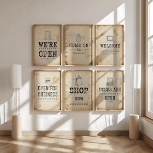 Op de afbeelding: Zes ingelijste borden in vintage-stijl met winkelgerelateerde zinnen. De borden bevatten "We're Open", "Come On In", "Welcome", "Open for Business", "Shop Now" en "Doors are Open". De borden hebben een verouderde uitstraling.