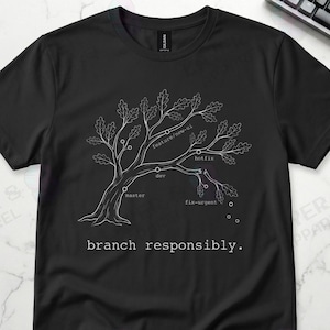 May include: Black t-shirt with a white tree graphic, featuring branches labeled 'master', 'dev', and 'feature/name-it'. The text 'branch responsibly' is printed below the tree. A keyboard and coffee mug are in the background.