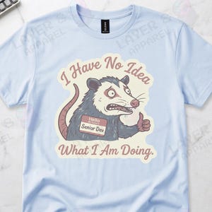 May include: Light blue t-shirt featuring a cartoon opossum with a name tag that reads "Hello Senior Dev." The opossum is giving a thumbs up, with the text "I Have No Idea What I Am Doing." printed above and below the opossum.