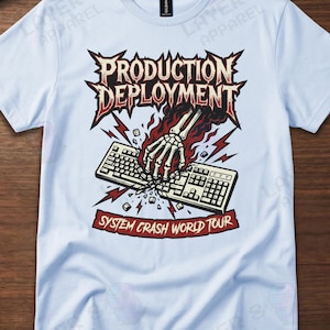 May include: Light blue t-shirt featuring a graphic design with the text "PRODUCTION DEPLOYMENT" above a skeletal hand breaking a keyboard. The text "SYSTEM CRASH WORLD TOUR" is below the keyboard. The design includes red and white accents.