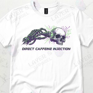 May include: White t-shirt featuring a graphic design. The design includes a skull being injected with a green and purple energy source by a robotic arm. The text "DIRECT CAFFEINE INJECTION" is below the graphic.