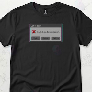 May include: Black t-shirt featuring a pixelated computer error message that reads "Life.exe Task Failed Successfully." The design includes a red "X" and buttons labeled "Panic," "Ignore," and "Reboot."