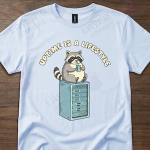 May include: Light blue t-shirt featuring a cartoon raccoon holding a cup, sitting atop a server rack. The text "Uptime is a lifestyle" arches above the raccoon. The shirt is a short-sleeved crew neck.