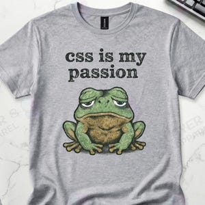 May include: A gray t-shirt with the text "css is my passion" above a cartoon frog with a grumpy expression. The frog is green and brown. The shirt is on a white surface with a keyboard and a black mug.