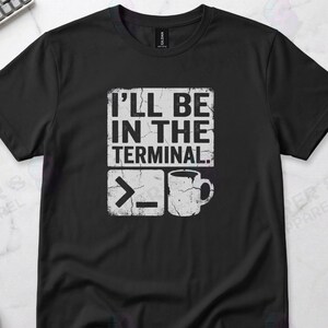 May include: Black t-shirt with the text "I'LL BE IN THE TERMINAL." in a white, distressed font. Below the text are a command prompt symbol and a coffee mug graphic. A casual tee for tech enthusiasts.