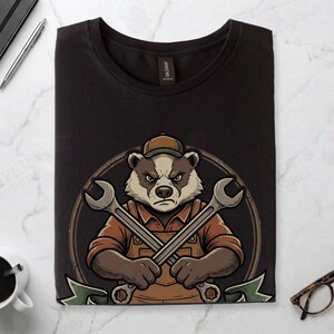 Black Gildan 64000 t-shirt featuring a vintage mechanic mascot illustration of a grumpy badger wearing overalls and holding two crossed wrenches above the text "FULL-STACK BADGER" on a green banner.