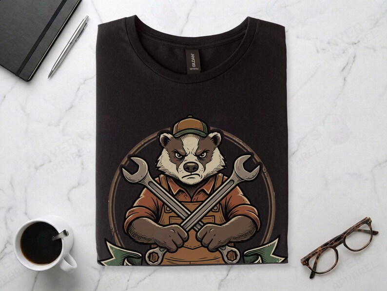 Black Gildan 64000 t-shirt featuring a vintage mechanic mascot illustration of a grumpy badger wearing overalls and holding two crossed wrenches above the text "FULL-STACK BADGER" on a green banner.