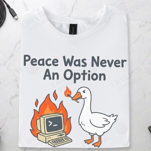 White Gildan t-shirt featuring a cartoon white goose holding a lit match next to a burning retro computer, with text "Peace Was Never An Option" and the Linux command "sudo rm -rf /".
