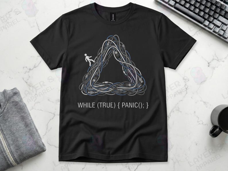 Black Gildan 64000 t-shirt featuring a Penrose impossible triangle made of tangled grey and white computer cables with a small white stick figure walking on it, above the white code text "WHILE (TRUE) { PANIC(); }".