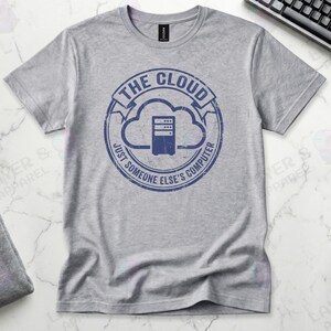 Sports grey t-shirt displaying a blue distressed stamp graphic. Text reads "THE CLOUD JUST SOMEONE ELSE'S COMPUTER" surrounding a server rack icon inside a cloud outline. A cynical technical definition of cloud infrastructure. Grunge vintage style.