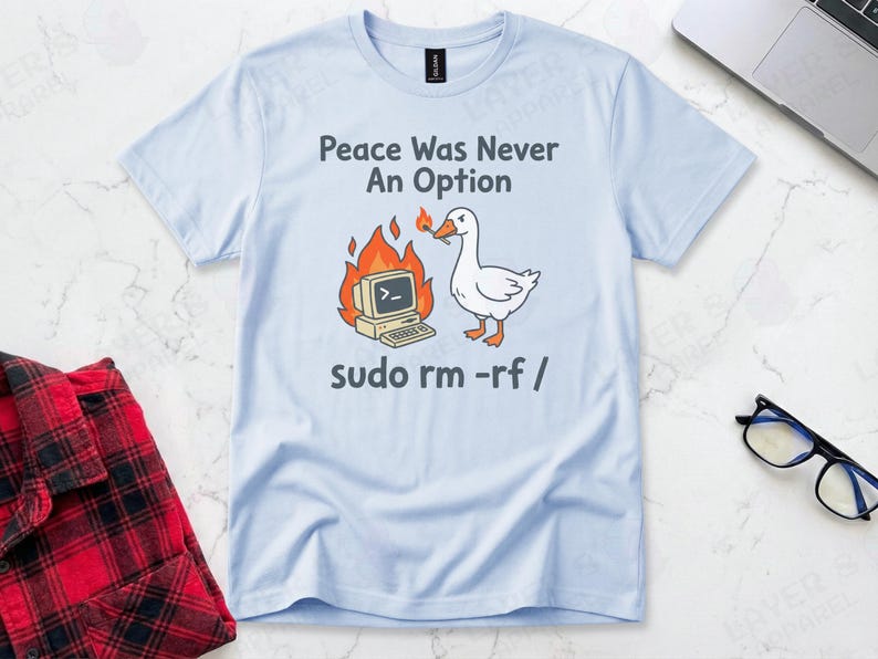 Light blue Gildan t-shirt featuring a cartoon white goose holding a lit match next to a burning retro computer, with text "Peace Was Never An Option" and the Linux command "sudo rm -rf /".