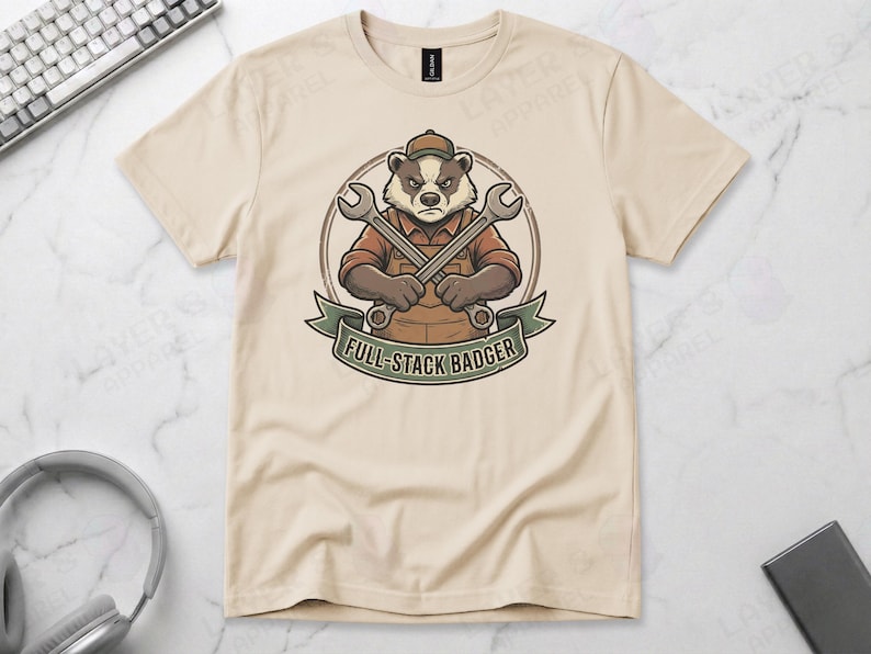 Sand beige Gildan 64000 t-shirt featuring a vintage mechanic mascot illustration of a grumpy badger wearing overalls and holding two crossed wrenches above the text "FULL-STACK BADGER" on a green banner.