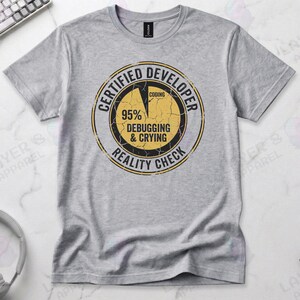 Sports grey t-shirt displaying a distressed circular badge graphic. Text reads "CERTIFIED DEVELOPER REALITY CHECK" surrounding a pie chart where 95% is labeled "DEBUGGING & CRYING" and a tiny sliver is "CODING". A humorous grunge-style statistic about the coding profession.