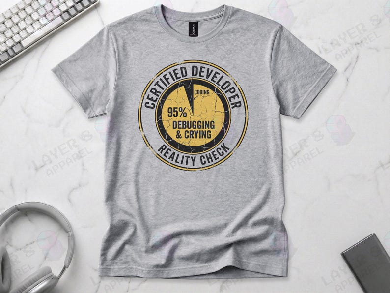 Sports grey t-shirt displaying a distressed circular badge graphic. Text reads "CERTIFIED DEVELOPER REALITY CHECK" surrounding a pie chart where 95% is labeled "DEBUGGING & CRYING" and a tiny sliver is "CODING". A humorous grunge-style statistic about the coding profession.