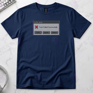 Navy blue t-shirt displaying a retro computer error window graphic with a glitch effect. Title bar reads "Life.exe" with a red X icon. Message reads "Task Failed Successfully." Buttons options are "Panic", "Ignore", and "Reboot." A humorous meme referencing contradictory system errors and existential anxiety.
