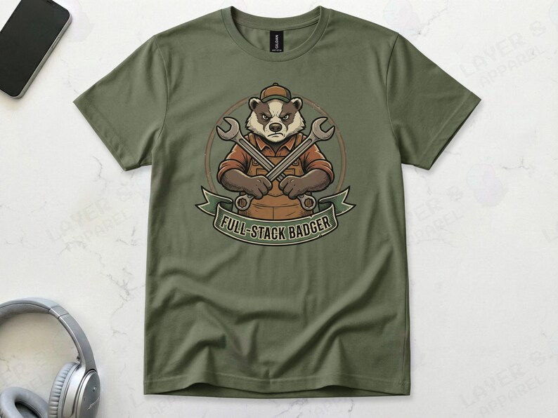 Military green Gildan 64000 t-shirt featuring a vintage mechanic mascot illustration of a grumpy badger wearing overalls and holding two crossed wrenches above the text "FULL-STACK BADGER" on a green banner.