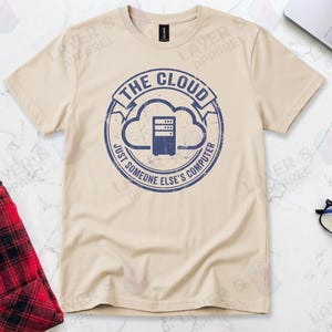 Sand t-shirt displaying a blue distressed stamp graphic. Text reads "THE CLOUD JUST SOMEONE ELSE'S COMPUTER" surrounding a server rack icon inside a cloud outline. A cynical technical definition of cloud infrastructure. Grunge vintage style.
