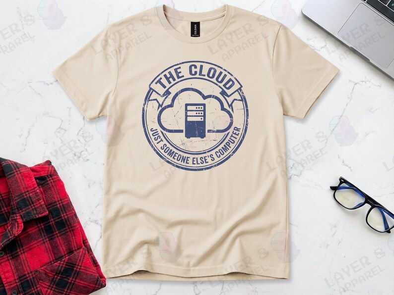 Sand t-shirt displaying a blue distressed stamp graphic. Text reads "THE CLOUD JUST SOMEONE ELSE'S COMPUTER" surrounding a server rack icon inside a cloud outline. A cynical technical definition of cloud infrastructure. Grunge vintage style.
