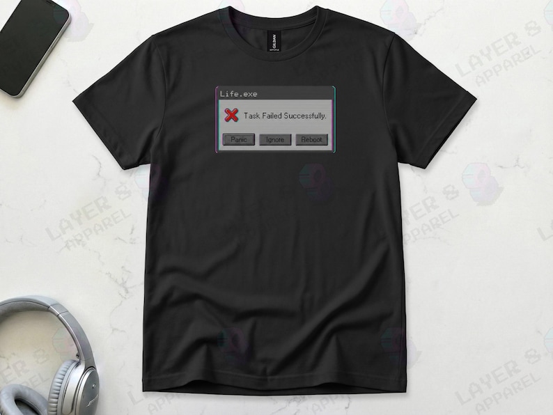 Black t-shirt displaying a retro computer error window graphic with a glitch effect. Title bar reads "Life.exe" with a red X icon. Message reads "Task Failed Successfully." Buttons options are "Panic", "Ignore", and "Reboot." A humorous meme referencing contradictory system errors and existential anxiety.