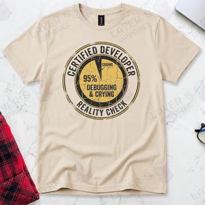 Sand t-shirt displaying a distressed circular badge graphic. Text reads "CERTIFIED DEVELOPER REALITY CHECK" surrounding a pie chart where 95% is labeled "DEBUGGING & CRYING" and a tiny sliver is "CODING". A humorous grunge-style statistic about the coding profession.