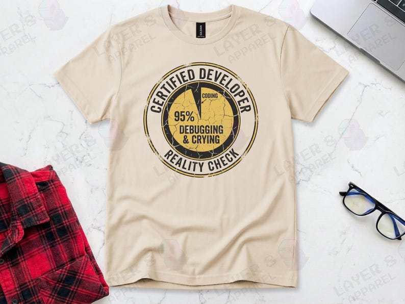 Sand t-shirt displaying a distressed circular badge graphic. Text reads "CERTIFIED DEVELOPER REALITY CHECK" surrounding a pie chart where 95% is labeled "DEBUGGING & CRYING" and a tiny sliver is "CODING". A humorous grunge-style statistic about the coding profession.
