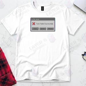 White t-shirt displaying a retro computer error window graphic with a glitch effect. Title bar reads "Life.exe" with a red X icon. Message reads "Task Failed Successfully." Buttons options are "Panic", "Ignore", and "Reboot." A humorous meme referencing contradictory system errors and existential anxiety.