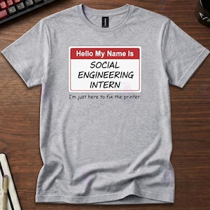 May include: A heather gray t-shirt with a name tag that reads "Hello My Name Is SOCIAL ENGINEERING INTERN". Below the name tag, the text reads "I'm just here to fix the printer."