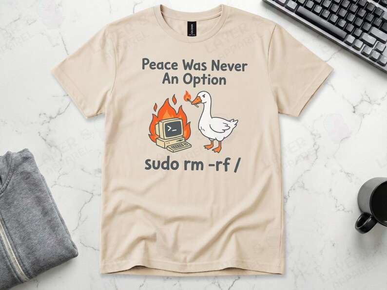 Sand Gildan t-shirt featuring a cartoon white goose holding a lit match next to a burning retro computer, with text "Peace Was Never An Option" and the Linux command "sudo rm -rf /".