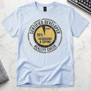 Light blue t-shirt displaying a distressed circular badge graphic. Text reads "CERTIFIED DEVELOPER REALITY CHECK" surrounding a pie chart where 95% is labeled "DEBUGGING & CRYING" and a tiny sliver is "CODING". A humorous grunge-style statistic about the coding profession.