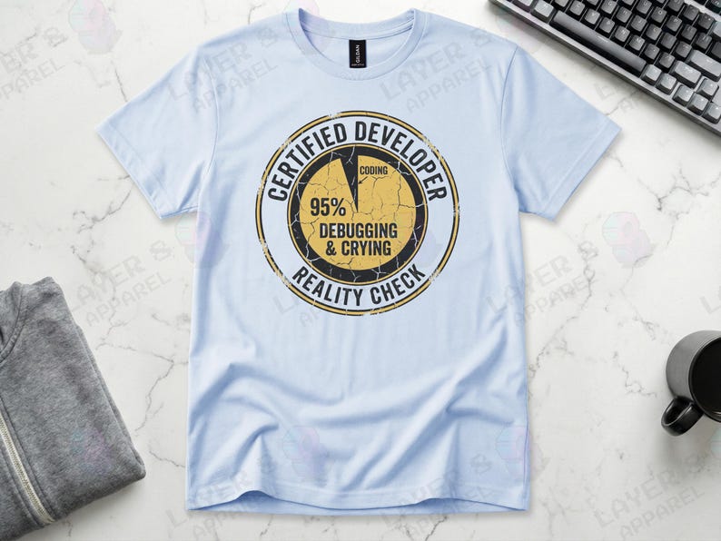 Light blue t-shirt displaying a distressed circular badge graphic. Text reads "CERTIFIED DEVELOPER REALITY CHECK" surrounding a pie chart where 95% is labeled "DEBUGGING & CRYING" and a tiny sliver is "CODING". A humorous grunge-style statistic about the coding profession.