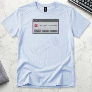 Light blue t-shirt displaying a retro computer error window graphic with a glitch effect. Title bar reads "Life.exe" with a red X icon. Message reads "Task Failed Successfully." Buttons options are "Panic", "Ignore", and "Reboot." A humorous meme referencing contradictory system errors and existential anxiety.