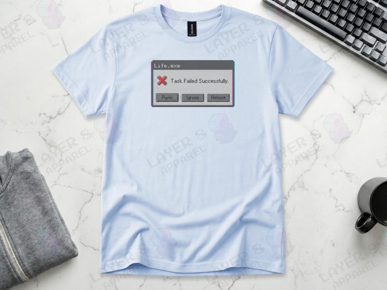 Light blue t-shirt displaying a retro computer error window graphic with a glitch effect. Title bar reads "Life.exe" with a red X icon. Message reads "Task Failed Successfully." Buttons options are "Panic", "Ignore", and "Reboot." A humorous meme referencing contradictory system errors and existential anxiety.