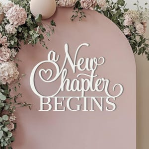 Peut inclure: Panneau en bois blanc avec l'inscription "a New Chapter BEGINS" en écriture élégante. Le panneau est placé sur un fond rose tendre, orné de compositions florales et d'une sphère décorative, parfait pour une célébration ou un événement.