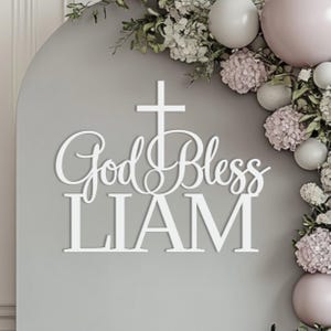 Op de afbeelding: Wit houten bord met de tekst "God Bless LIAM" in sierlijk lettertype, bekroond met een kruis. Het bord staat tegen een grijze achtergrond, versierd met bloemen en ballonnen, ideaal voor een doop of communie.