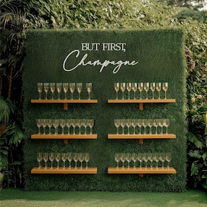 Può includere: Un muro verde con la scritta "BUT FIRST, Champagne" in bianco. Mensole in legno sostengono file di calici di champagne, ciascuno riempito con un liquido dorato. L'esposizione è all'aperto, circondata dal verde, creando una presentazione elegante.