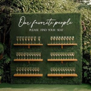 Könnte beinhalten: Eine grüne Wand mit dem weißen Schriftzug "Our favorite people PLEASE FIND YOUR SEAT". Holzregale halten Reihen von Sektgläsern, die eine Sitzordnung für eine Hochzeit oder Veranstaltung bilden.
