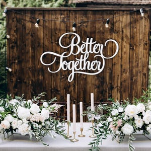 May include: Wedding reception decor with a wooden backdrop and the words "Better Together" in white script. The table is decorated with white flowers, greenery, candles, and glasses. String lights hang above.