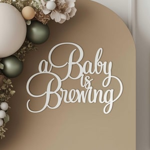 May include: White cursive text on a brown background reads "a Baby is Brewing." The text is part of a decorative sign, with balloons and floral accents in the upper left corner. The balloons are white, green, and tan.
