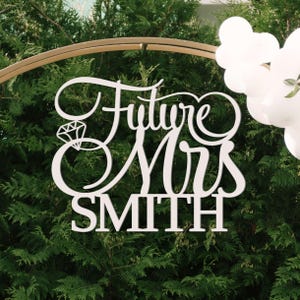Può includere: Cartello in legno bianco con la scritta "Future Mrs SMITH" in elegante corsivo, con un design ad anello con diamante. Il cartello è posto su uno sfondo di fogliame verde e palloncini bianchi, ideale per un matrimonio o un addio al nubilato.