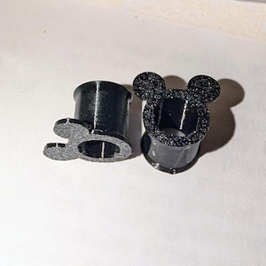 Puede incluir: Dos dilatadores de oreja negros con un diseño de cabeza de Mickey Mouse. Uno está tumbado de lado, el otro está de pie. Los dilatadores tienen una textura brillante en la cabeza de Mickey Mouse y alrededor del borde.