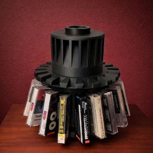 May include: A black cassette tape holder with a circular design, holding several cassette tapes. The holder has a cylindrical top with vertical slots and a gear-like base. The tapes are visible, with some text on the labels.