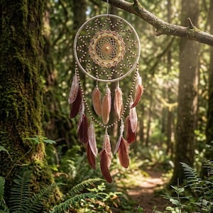 May include: A handcrafted dreamcatcher with a circular woven web design, adorned with brown feathers and wooden beads. The dreamcatcher hangs from a tree branch in a lush forest setting, with sunlight filtering through the trees.